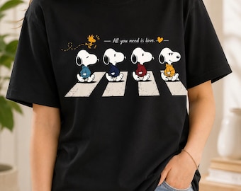 May include: A black short-sleeved t-shirt featuring a graphic of four Snoopy characters in colored hoodies walking across a crosswalk. Above them, the text reads All you need is love with a small yellow heart and a flying Woodstock character.