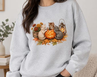 May include: A light gray crewneck sweatshirt featuring a graphic print of three kittens surrounding a glowing jack-o'-lantern, a wooden crate of orange flowers, a vintage-style lantern, and scattered autumn leaves on the front center.