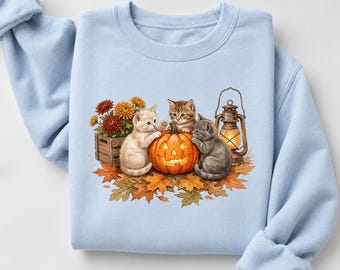 May include: A light blue crewneck sweatshirt featuring a graphic print of three kittens surrounding a glowing jack-o'-lantern. The scene includes a wooden crate of orange chrysanthemums, a lit vintage-style lantern, and scattered autumn leaves.