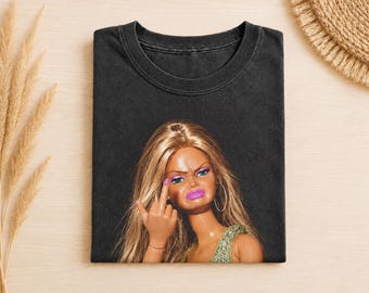 May include: A charcoal black t-shirt featuring a graphic print of a blonde doll with long hair, wearing hoop earrings and a green top. The doll has a frowning expression, bright pink lipstick, and is holding up a middle finger gesture.
