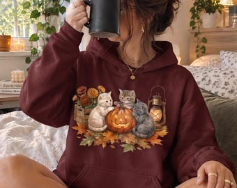 May include: A dark maroon hooded sweatshirt featuring a graphic print of three kittens sitting around a glowing jack-o'-lantern, surrounded by autumn leaves, a wooden crate of orange flowers, and a vintage-style lantern with a warm light.