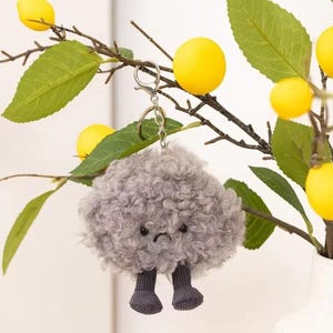 Puede incluir: Un llavero de felpa gris con forma de nube con una cara triste y piernas grises adjuntas. El llavero cuelga de un cierre plateado. El fondo incluye una rama con limones amarillos y hojas verdes.