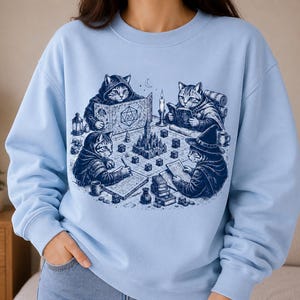 Op de afbeelding: Een lichtblauw sweatshirt met ronde hals en een donkerblauwe grafische print van vier katten die een tabletop rollenspel spelen. De illustratie toont de katten met dobbelstenen, boeken, een kasteelmodel en een scherm met een 20-zijdige dobbelsteen.