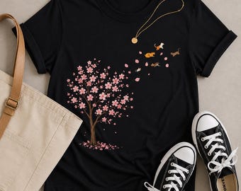 May include: A black short-sleeved t-shirt featuring a graphic of a cherry blossom tree with pink flowers. Small cats in various colors are depicted as if they are being carried away by the wind along with the falling pink flower petals.