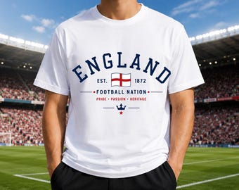 May include: A white short-sleeved t-shirt featuring the text ENGLAND in dark blue, the English flag, and the text EST. 1872. Below this, the text FOOTBALL NATION, PRIDE PASSION HERITAGE, and a small blue crown icon with a red star are printed in blue and red.