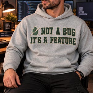 Könnte beinhalten: Ein graumelierter Kapuzenpullover mit einer Grafik eines grünen Käfers, der mit einer diagonalen Linie durchgestrichen ist, sowie dem dunkelgrünen Text: NOT A BUG IT'S A FEATURE. Der Hoodie verfügt über eine Kängurutasche und lange Ärmel.