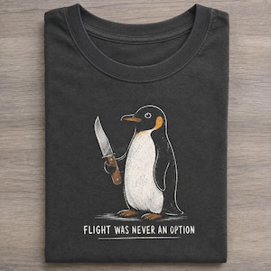 May include: A charcoal grey crew neck t-shirt featuring a graphic illustration of a penguin holding a knife. Below the penguin, the text reads FLIGHT WAS NEVER AN OPTION in white capital letters. The design uses black, white, and orange tones.