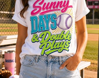 May include: A white short-sleeve t-shirt featuring the text Sunny DAYS & Double Plays in pink, blue, and lime green, accompanied by a baseball graphic with yellow sunburst rays. The shirt is paired with light blue distressed denim shorts.