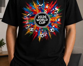 May include: A black short-sleeved t-shirt featuring a central circular graphic with the text 2026 WORLD CUP. The design includes a colorful paint splatter effect, various national flags, and several soccer balls arranged around the center text.