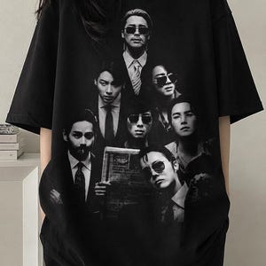 May include: Black short-sleeved t-shirt featuring a grayscale image of a group of people in suits and sunglasses. The image is centered on the front of the shirt.