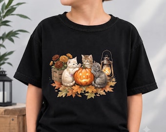 May include: A black t-shirt featuring a graphic print of three kittens gathered around a glowing jack-o'-lantern. The scene includes a wooden crate with orange flowers, scattered autumn leaves, and a lit vintage-style lantern on the side.