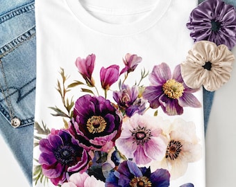 May include: A white t-shirt featuring a vibrant floral print of purple, violet, and cream-colored anemone flowers. The design includes detailed petals and dark centers. Two fabric hair scrunchies in purple and beige are placed near the shirt.