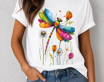 May include: A white short-sleeved t-shirt featuring a vibrant, watercolor-style graphic of a large dragonfly with multicolored wings in shades of blue, yellow, orange, and pink, surrounded by various small wildflowers and stems.