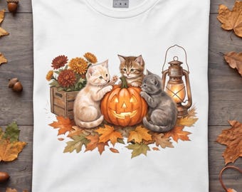 May include: A white t-shirt featuring a graphic of three kittens surrounding a glowing jack-o'-lantern, a wooden crate filled with orange and red chrysanthemums, and a lit lantern, all resting on a bed of fallen autumn leaves.