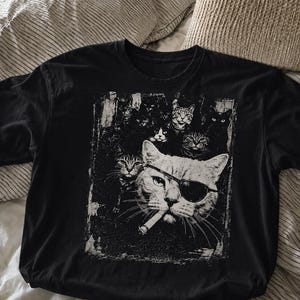 May include: Black t-shirt featuring a distressed graphic of a cat wearing an eye patch and sunglasses, smoking a cigarette, surrounded by other cats. The design is in shades of white and gray.