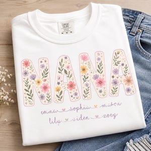 May include: A white t-shirt featuring the word NANA in large block letters filled with a colorful floral pattern of pink, purple, and yellow flowers. Below the word, two lines of cursive text list names separated by small heart icons.