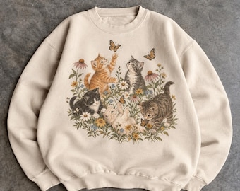May include: A beige crewneck sweatshirt featuring a central graphic illustration of four kittens playing among colorful wildflowers and butterflies. The kittens are depicted in orange tabby, grey tabby, black and white, and cream fur patterns.