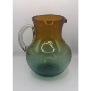 May include: A two-toned glass pitcher with a handle and a spout. The top portion is amber, transitioning to a green base. The pitcher is likely used for serving beverages.