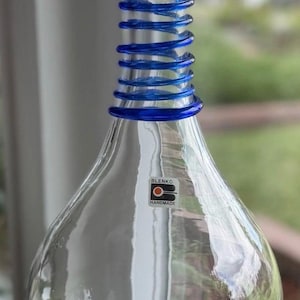 Pode incluir: Um decantador de vidro transparente com uma base larga e arredondada e um pescoço alto e fino. O pescoço apresenta uma espiral de vidro azul enrolada à volta. Um pequeno autocolante retangular no corpo diz BLENKO HANDMADE com um logótipo.