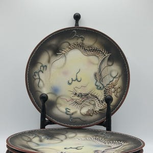 Op de afbeelding: Een set decoratieve keramische borden met een drakenontwerp. De borden hebben een donkerbruine rand en een crèmekleurig midden met een gedetailleerde drakenillustratie in bruin- en wittinten. De borden worden gepresenteerd op een zwarte metalen standaard.