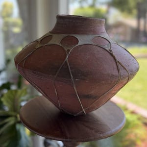 May include: A terracotta-colored ceramic vessel with a wide, angular body and a narrow neck. The surface features a decorative, light-colored web-like pattern of interconnected circles and lines that wrap around the midsection of the pot.