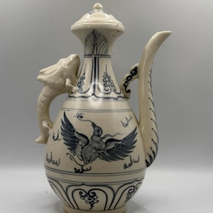 May include: A cream-colored ceramic pitcher with a lid, featuring blue painted bird and floral designs. The pitcher has a curved spout and a handle shaped like a dragon. Decorative Asian-inspired art.