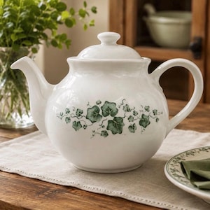 May include: A white ceramic teapot featuring a rounded body, a matching lid with a knob handle, and a curved spout. The exterior is decorated with a horizontal band of green ivy leaves and delicate vine tendrils wrapping around the center.