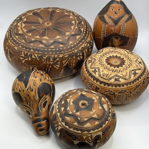 Può includere: Cinque zucche marroni con intricati disegni intagliati. Le zucche variano per forma e dimensione, con motivi dettagliati tra cui forme geometriche e figure. I colori vanno dal beige chiaro al marrone scuro.