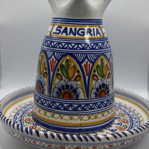 May include: A white ceramic sangria pitcher with a matching plate. The pitcher has a blue and yellow floral design with the word "SANGIRA" in blue letters. The plate has a matching design.