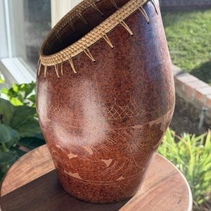 May include: A handcrafted, brown gourd-shaped basket with etched designs. The basket has a woven rim and is set on a wooden stand. The gourd has a natural, organic look, with a slightly tilted opening.