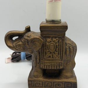 May include: A brown elephant-shaped lamp with a decorative design. The lamp has a rectangular base and a square top that holds the light fixture. The lamp has a label that reads "WARNING: TO REDUCE THE RISK OF FIRE 40 WATT TYPE LAMP MAXIMUM."
