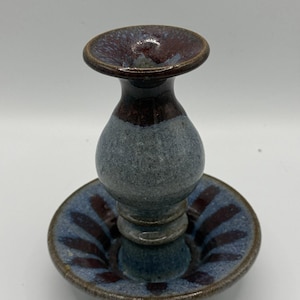 May include: A handcrafted ceramic candlestick holder. The holder features a dark blue and burgundy color scheme. The base is a shallow dish with burgundy stripes, and the candlestick has a bulbous body and a flared top.