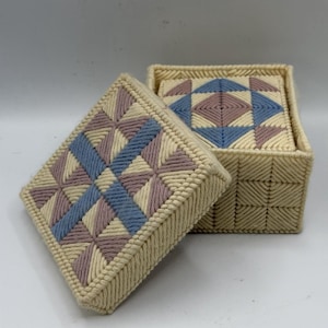 May include: A square, beige yarn box with a lid, featuring a geometric pattern in blue, lavender, and beige. The lid is open, revealing a matching pattern inside. The box and lid are made of yarn.