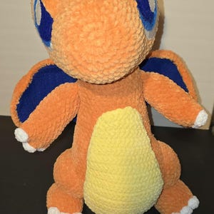 May include: A plush orange creature with blue accents and large blue eyes. The stuffed animal has a yellow belly and white accents on its feet and hands. The toy is handmade and appears to be crocheted.