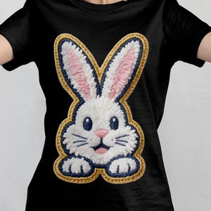 Könnte beinhalten: Schwarzes T-Shirt mit einem großen, gestickten Hasenmuster. Der Hase hat weißes Fell, rosa Innenohren, blaue Augen und einen lächelnden Mund. Das Design ist gold und blau umrandet.