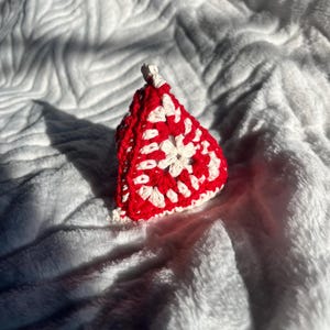 以下が含まれることがあります： 手作りの赤と白のかぎ針編みオーナメント。オーナメントは三角形で、中央に白い花の模様があります。かぎ針編みの作品が見え、オーナメントは柔らかく白い質感の表面に置かれています。