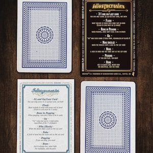 May include: Four playing cards and two rule cards are displayed on a wooden surface. The playing cards have a blue and white geometric design on the back. The rule cards have text that describes the rules of the card game "Idiosyncrasies".