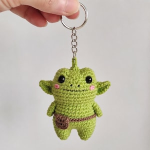 Op de afbeelding: Een lichtgroene gehaakte sleutelhanger in de vorm van een wezen met grote zwarte ogen, kleine roze wangen en een bruine riem met een klein tasje. Het figuurtje heeft puntige oren en hangt aan een zilverkleurige metalen ring en ketting.