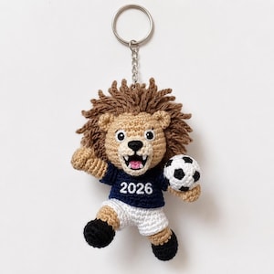 Könnte beinhalten: Gehäkelter Löwen-Schlüsselanhänger mit Fußball. Der Löwe hat eine braune Mähne und einen beigefarbenen Körper und trägt ein marineblaues Trikot mit der Zahl 2026. Der Schlüsselanhänger hat einen silbernen Ring.