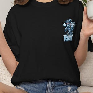 May include: Black t-shirt featuring a cartoon yeti graphic with the word "ICEMAN" in blue and white. The yeti is depicted in shades of blue and white, with a frosty appearance. The shirt is a casual wear item.