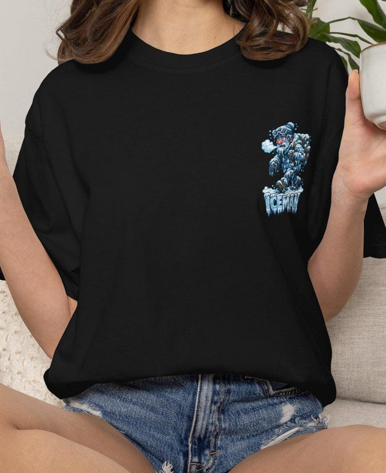 May include: Black t-shirt featuring a cartoon yeti graphic with the word "ICEMAN" in blue and white. The yeti is depicted in shades of blue and white, with a frosty appearance. The shirt is a casual wear item.