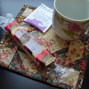 May include: A white teacup with pink floral designs sits on a floral quilted coaster. A package of Melinda's Biscotti, labeled "Chocolate Cranberry", and a Tazo tea bag are also present, suggesting a cozy tea break.