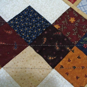 May include: A quilt made of various fabrics with different patterns and colors. The quilt is made in a diamond pattern with a white background. The fabrics are brown, blue, orange, and beige.
