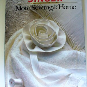 May include: A white book with the title "More Sewing for the Home" and the word "SINGER" in red letters above the title. The book cover features a white fabric rose and a white towel.