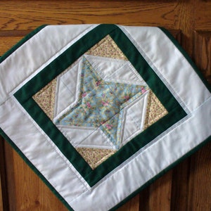 May include: A white quilted table runner with a green border and a star-shaped design in the center. The star is made of different fabrics with floral patterns in shades of blue, yellow, and pink.