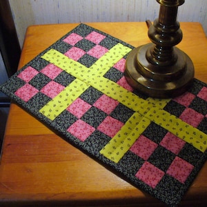 May include: A checkered hot pad with pink and black squares and yellow cross-shaped accents. The hot pad is made of fabric and has a floral pattern.