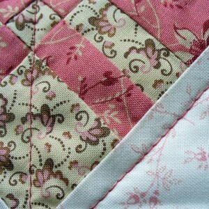 May include: A close-up of a quilt made with different patterned fabrics. The quilt is made with pink and white fabrics with floral patterns. The quilt is sewn together with a white thread.