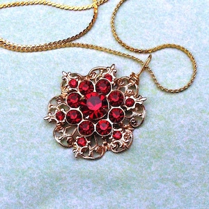 May include: A gold-toned necklace featuring a pendant with a floral design. The pendant is adorned with numerous red gemstones, with a larger central stone. The chain is a delicate, woven style.