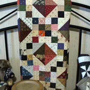 May include: A patchwork quilt featuring a variety of fabric squares in shades of red, blue, green, brown, and cream. The design incorporates geometric patterns, including large triangles and smaller squares, with visible stitching throughout the fabric.