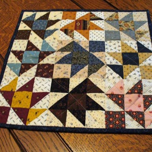 May include: A square, handmade quilt with a dark blue border. The quilt features a patchwork design with various geometric shapes and patterns in shades of beige, brown, blue, and yellow. The quilt is likely made of cotton.